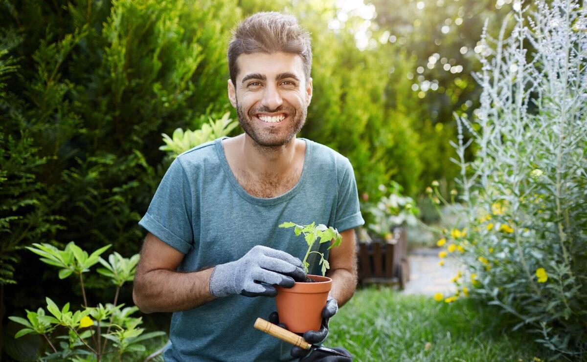 Outdoors portrait of young good-looking caucasian bearded man in blue shirt and gloves smiling in camera, holding pot with flower in hands working in garden.