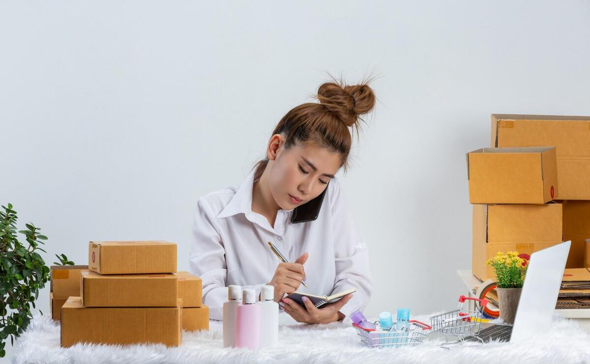 Woman jotting notes while on phone amidst parcels and cosmetics on a white backdrop.