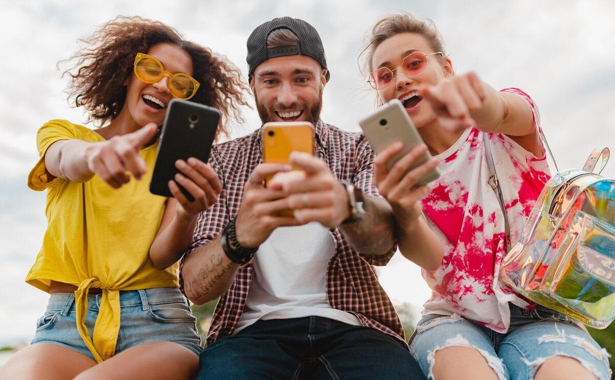 Happy young company of smiling friends sitting park using smartphones