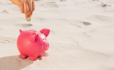 A hand placing a coin into a vibrant pink piggy bank on a sunlit, sandy beach.