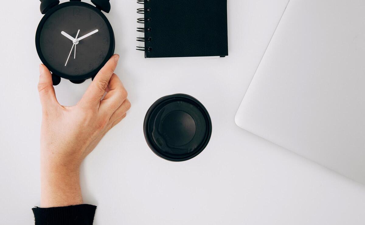 Close-up of a person's hand holding black alarm clock; spiral notepad; laptop and takeaway coffee cup on white desk