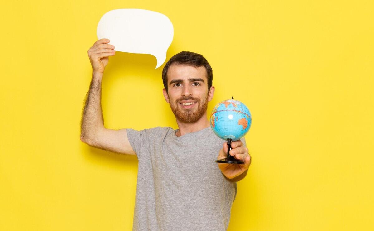 A front view young male in grey t-shirt holding white sign and little globe on the yellow wall man color model emotion clothes