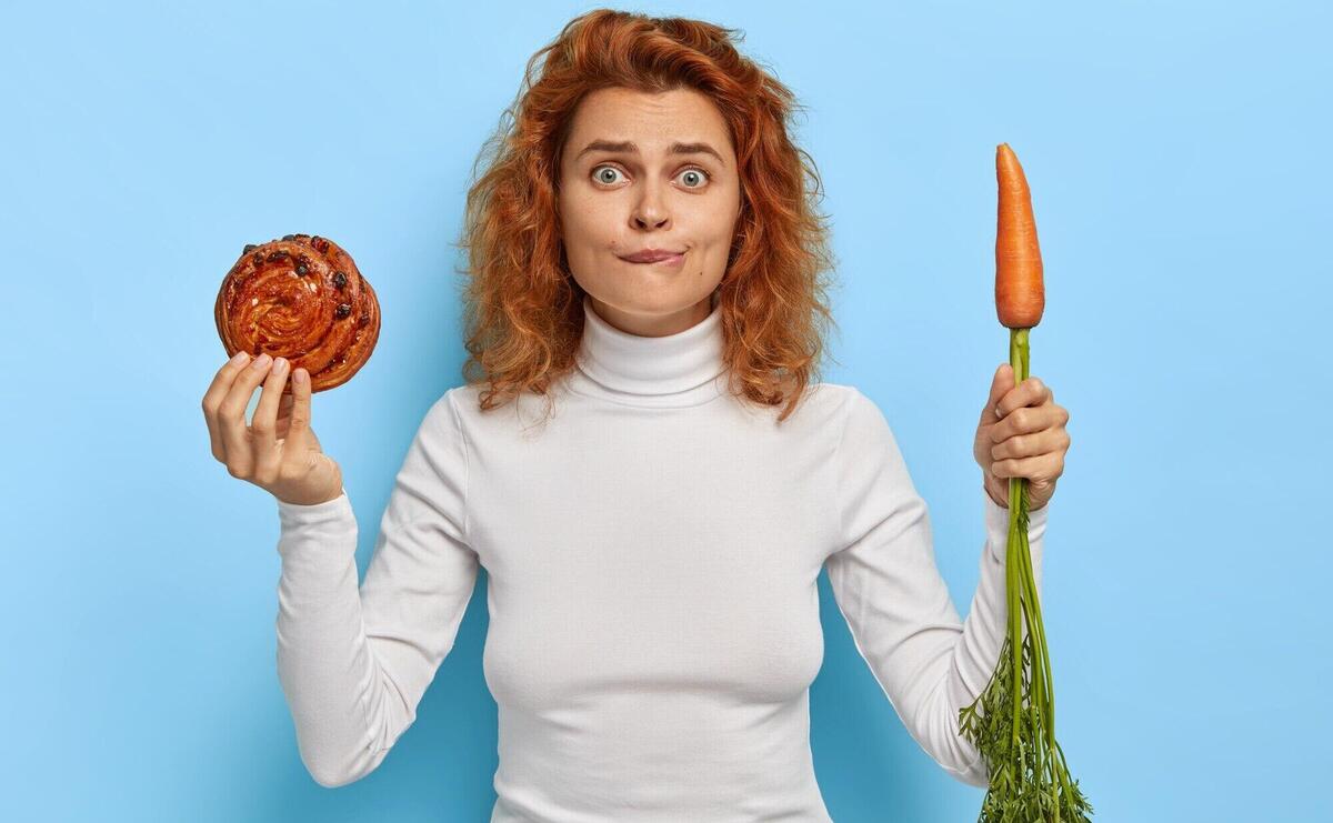 People, nutrition, dieting and junk food concept. embarrassed redhead woman holds fresh tasty bun and carrot, chooses between vegetable and confectionery, wears white turtleneck, stands indoor