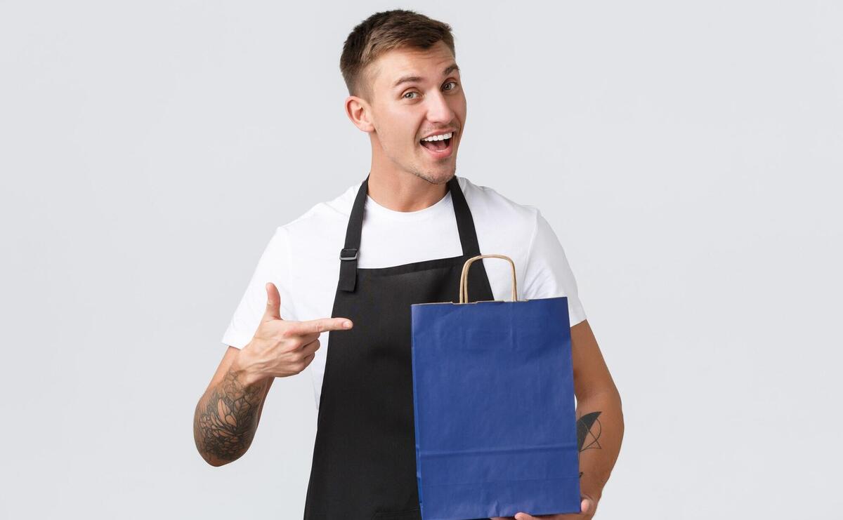 Enthusiastic friendly salesman in black apron, pointing finger at eco-bag.