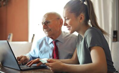Young woman in casual clothes helping senior man in formal shirt with paying credit card in Internet using laptop while sitting at table