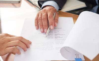 Close-up of hands reviewing a document with a pen, with books and a clipboard nearby.